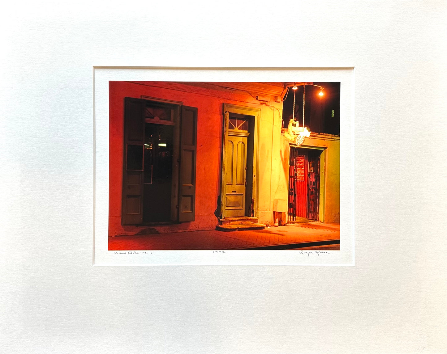 This photograph was taken in the French Quarter of New Orleans.  The buildings have large shutters around one of the doors and heavy wood around the other  two doors. The lighting on the street produces a heated red shaded on the left side with a very warm feeling of yellow-green on the right.  It looks like the evening is about to start but the people have not arrived yet.